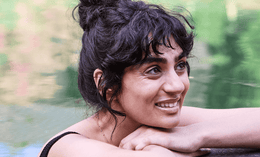 A woman with dark curly hair in a messy bun smiles while resting her arms on a stone ledge by a body of water, with a blurred green background.