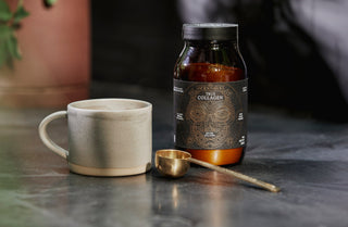 A ceramic cup, a brass measuring spoon, and a brown jar labeled Aventurine True Collagen are arranged on a dark surface with a potted plant blurred in the background.