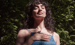 A woman with long dark hair stands outdoors in front of green foliage, eyes closed and one hand resting on her chest, appearing peaceful and content as sunlight falls on her face.