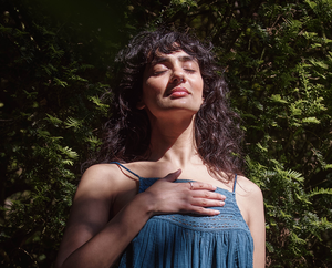 A woman with long dark hair stands outdoors in front of green foliage, eyes closed and one hand resting on her chest, appearing peaceful and content as sunlight falls on her face.