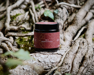 A jar labeled True Ashwagandha sits on large, textured tree roots surrounded by greenery and natural light.