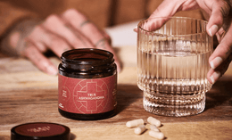 A person’s hand holds a glass of water next to a jar labeled “True Ashwagandha” with its lid off and four capsules lying on a wooden surface.