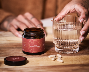A person’s hand holds a glass of water next to a jar labeled “True Ashwagandha” with its lid off and four capsules lying on a wooden surface.