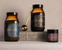 Three jars of collagen supplements with intricate labels are displayed on a stone block. A gold measuring spoon labeled ANCIENTBRAVE rests between the two larger jars. Background is neutral beige.