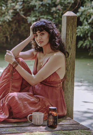 A woman in a flowing rose-colored dress sits on a wooden dock by a pond, resting her head on her hand. A ceramic mug and a brown bottle are placed beside her, with lush greenery in the background.