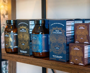 Bottles and boxes of Ancestral Wild Collagen supplements are displayed on a wooden shelf. The packaging features intricate gold and blue designs with product information and branding clearly visible.