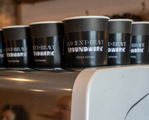 A row of black coffee cups labeled Ancient + Brave Groundwork Abbot Kinney is lined up on top of a white espresso machine in a coffee shop.