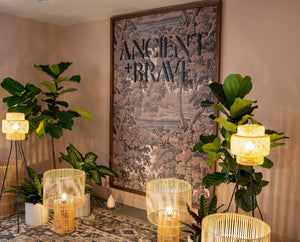 A cozy room with large green plants, wicker floor lamps, a patterned rug, and a large framed artwork on the wall that reads ANCIENT + BRAVE above a nature scene.