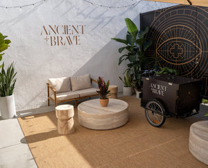 A modern outdoor lounge with a white sofa, round stone tables, potted plants, a rug, and a black cart labeled Ancient + Brave against a white wall with geometric and eye artwork.