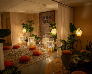 A cozy, softly-lit room with orange floor cushions arranged in a circle, surrounded by green plants, woven lamps, sheer curtains, and a patterned rug, creating a warm, inviting atmosphere for relaxation or meditation.