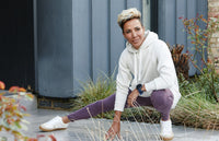 A person in a white hoodie, purple leggings, and white sneakers crouches outdoors on a paved area, smiling and looking to the side, with modern building panels and plants in the background.