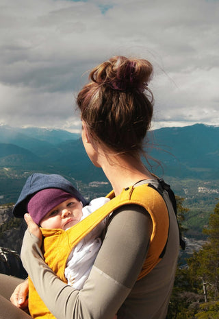 A woman with a bun holds a baby in a yellow carrier while sitting on a mountainside, overlooking a scenic landscape of mountains, trees, and cloudy sky.