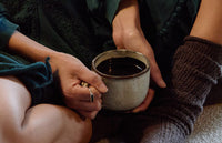 A person wearing dark socks and a green dress holds a ceramic mug filled with a dark beverage, sitting with legs crossed on a cozy surface.