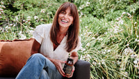A woman with long brown hair, wearing a white blouse and blue jeans, sits on an outdoor couch, smiling and holding a bottle, with green plants and white flowers in the background.