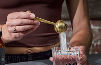 A person in a brown crop top pours a heaping spoonful of white powder into a glass of liquid, likely preparing a drink. Their hand, wearing a ring, holds a gold-colored spoon above the glass.