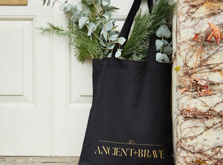 A black tote bag with ANCIENT + BRAVE in gold text leans against a door, filled with green foliage and eucalyptus branches.