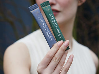 A person in a sleeveless top holds up two Ancient + Brave supplement sachets, one dark blue and one green, with the front labels facing the camera. The background is blurred.