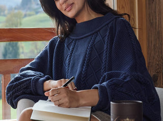 A woman in a blue sweater sits by a window, smiling and writing in a notebook. A mug and a tin container are on the table beside her. Green hills are visible through the window in the background.