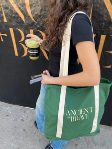 A person with long wavy hair holds two limited edition snack bars and a green drink, carrying the Limited Edition Tote Bag from Ancient + Brave US. They wear a black shirt and blue jeans.