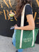 A person with long wavy hair holds two limited edition snack bars and a green drink, carrying the Limited Edition Tote Bag from Ancient + Brave US. They wear a black shirt and blue jeans.
