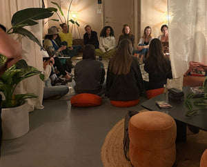 Group of people sitting on orange stools in a room with plants and a mirror.