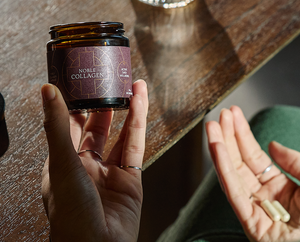 A person holds a jar labeled Noble Collagen in one hand and two capsules in the other, sitting at a wooden table in bright natural light.