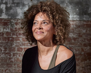 A woman with curly hair, wearing an off-the-shoulder black top and an olive green strap, smiles softly while sitting against a worn brick wall with a gray, weathered texture.
