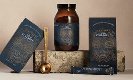 A jar, two boxes, a sachet, and a gold measuring spoon of Ancient + Brave Wild Collagen are arranged on a stone with a beige background. The packaging features intricate gold line art on dark blue.