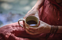 A person in a rust-colored dress sits outdoors, holding a ceramic mug filled with dark coffee in both hands. Sunlight creates soft patterns on the background.