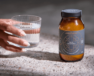 A hand with manicured nails holds a glass of water next to a brown jar labeled Ancient + Brave True Creatine+ Advanced Creatine Formula, both placed on a textured stone surface.