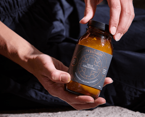 A person holds a brown glass jar labeled Ancient+Brave True Creatine+ Advanced Creatine Formula with both hands, against a background of dark clothing and a light surface.