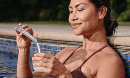 A woman in a pool, wearing a maroon swimsuit, stirs a powdered supplement into a glass of water with a smile, holding a blue packet next to the pool edge.