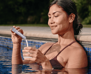 A woman in a pool, wearing a maroon swimsuit, stirs a powdered supplement into a glass of water with a smile, holding a blue packet next to the pool edge.