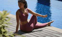 A woman in a mauve athletic outfit sits on a wooden deck by a pool, with one leg extended and the other bent, relaxing in the sun with her eyes closed.