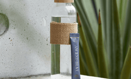 A clear glass water bottle with a tan woven sleeve and cap stands on a stone surface next to a blue Ancient + Brave drink sachet. Green, spiky plant leaves are blurred in the background.