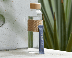 A clear glass water bottle with a tan woven sleeve and cap stands on a stone surface next to a blue Ancient + Brave drink sachet. Green, spiky plant leaves are blurred in the background.