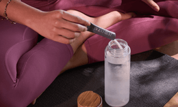 A person in mauve workout clothes sits cross-legged on a yoga mat, holding a stick labeled LIVELY VITAMIN CO. and pouring its contents into a clear water bottle with a bamboo lid beside it.