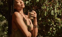 A woman with long braids stands outdoors in sunlight, eyes closed and smiling, holding a ceramic cup. She wears a sleeveless dress and bracelets, surrounded by green foliage.