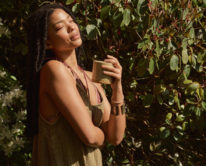 A woman with long braids stands outdoors in sunlight, eyes closed and smiling, holding a ceramic cup. She wears a sleeveless dress and bracelets, surrounded by green foliage.