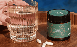 A hand holds a glass of water next to a jar labeled Triple Magnesium+ and three white capsules on a wooden tray, with a blue background.