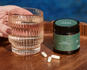 A hand holds a glass of water next to a jar labeled Triple Magnesium+ and three white capsules on a wooden tray, with a blue background.