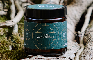 A dark jar labeled Triple Magnesium+ sits on a tree root outdoors, surrounded by moss and bark, promoting whole body health and daily ritual use.