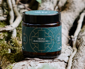 A dark jar labeled Triple Magnesium+ sits on a tree root outdoors, surrounded by moss and bark, promoting whole body health and daily ritual use.