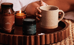 A hand holds a ceramic mug on a wooden tray with jars labeled True Nightcap and True Magnesium, a lit candle, and small stones, all set on a woven mat.