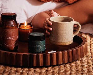 A hand holds a ceramic mug on a wooden tray with jars labeled True Nightcap and True Magnesium, a lit candle, and small stones, all set on a woven mat.