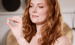 A woman with long red hair holds up a white capsule pill between her fingers, looking at it thoughtfully indoors.