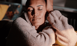 A person with a shaved head and closed eyes sits peacefully on a yellow couch, resting their head on one hand and wearing a soft, textured beige sweater. Sunlight gently illuminates their serene face.