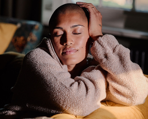 A person with a shaved head and closed eyes sits peacefully on a yellow couch, resting their head on one hand and wearing a soft, textured beige sweater. Sunlight gently illuminates their serene face.
