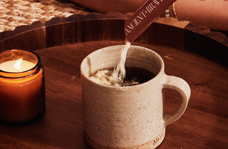 A hand pours powder from a sachet labeled ANCIENT + BRAVE into a beige ceramic mug filled with a dark beverage. A lit brown candle sits nearby on the wooden tray.