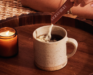 A hand pours powder from a sachet labeled ANCIENT + BRAVE into a beige ceramic mug filled with a dark beverage. A lit brown candle sits nearby on the wooden tray.